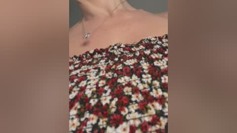 Media: A close-up video of a woman's neck and chest, wearing a strapless, floral-patterned dress with red, white, and black flowers. She has a silver necklace with a small pendant. The background is blurred, focusing on the dress's texture and her skin.