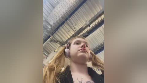 Media: Video of a young woman with fair skin and long blonde pigtails wearing headphones, standing in a warehouse with a corrugated metal roof.