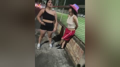 Media: Video of two women, one wearing a black one-shoulder top and shorts, the other in a white tank top and pink shorts, leaning against a chain-link fence.