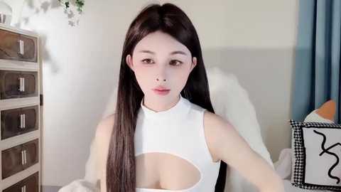 Media: Video of an East Asian woman with long, straight black hair and pale skin, wearing a white, sleeveless top with a keyhole cutout, sitting in a minimalist room with a white bed, wooden drawers, and a checkered box.