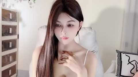 Media: Video of a young, fair-skinned Asian woman with long, straight brown hair, wearing a white spaghetti strap top, sitting on a white chair in a minimalist room with beige walls and a wooden dresser.