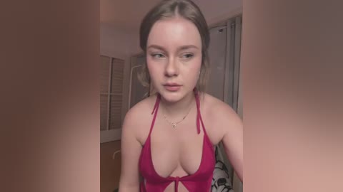 Media: Video of a young Caucasian woman with fair skin and long, light brown hair, wearing a red halter bikini top, looking contemplative in a dimly lit, simple bedroom.