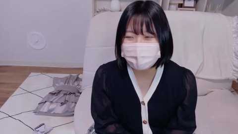 Media: Video of an Asian woman with short black hair, wearing a black cardigan and a white face mask, sitting on a white bedspread with a gray pattern, in a minimalist bedroom with light walls and wooden floor.