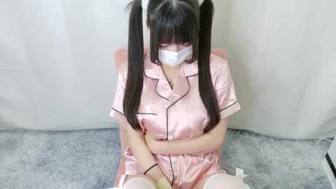 Media: A video of an East Asian woman with long black pigtails, wearing a pink satin nurse uniform, face mask, and sitting on a gray carpet.