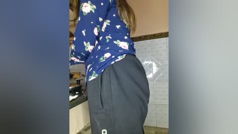 Media: Video of a woman with long brown hair wearing a blue floral top and black pants, standing in a kitchen with beige walls and tiled backsplash.