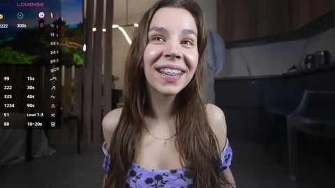 Media: Video of a smiling teenage girl with braces, long brown hair, in a purple off-shoulder top, indoors, kitchen background, digital overlay showing camera controls.