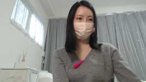 Media: Video of an East Asian woman with straight black hair, wearing a gray V-neck sweater, pink scarf, and a face mask, standing in a modern, bright room with white walls, a window, and gray curtains.