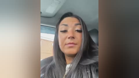 Media: A video of a middle-aged woman with medium-brown skin and long, straight, gray hair, wearing a white blouse, sitting in a car with a beige interior.