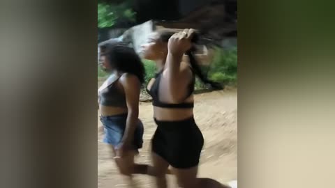 Media: Video of two women with medium skin tones, one with curly hair in a black crop top and shorts, the other with straight hair in a black sports bra and high-waisted shorts, standing on a dirt path in a lush, green forest setting.