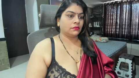 Media: Video of a South Asian woman with medium complexion and long black hair, wearing a red sari with a black lace bra. She sits on a gray chair in a simple room with a bed and window.