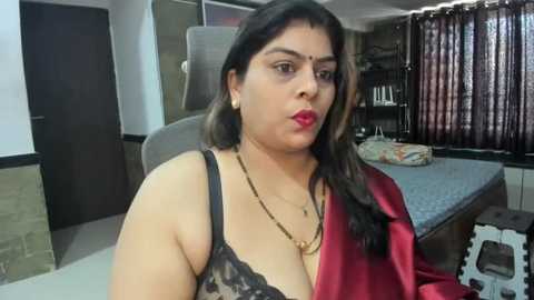 Media: Video of a South Asian woman with medium skin tone, wearing a black lace bra and red satin robe, sitting on a bed in a modern bedroom with grey curtains and wooden furniture.