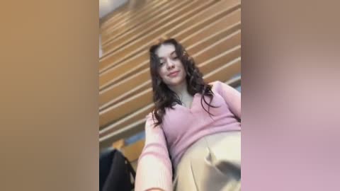 Media: Video of a young woman with long, wavy dark hair, wearing a pink sweater and beige pants, standing in a modern, wooden-paneled building with a blurred background.