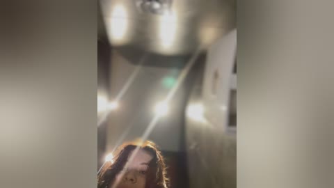 Media: A video shows a blurry, dimly lit hallway with a ceiling light fixture, blurred figures, and a partially visible person with shoulder-length hair. The image has a soft focus effect.