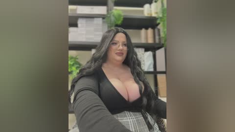 Media: Video of a curvy, light-skinned woman with long black hair, wearing a low-cut black top, sitting in an office with shelves of books and plants in the background.