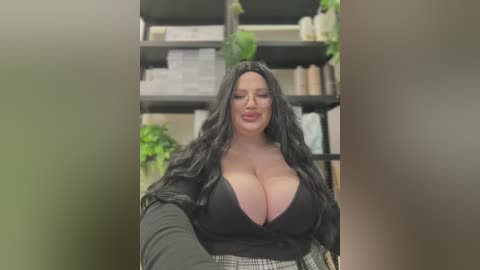 Media: Video of a curvy, dark-haired woman with long wavy hair, wearing a plunging black top revealing large breasts, standing in front of a green plant-filled bookshelf.