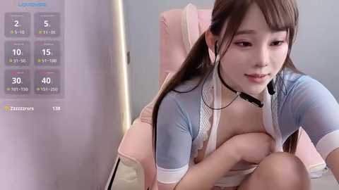 Media: Video of a young Asian woman with long brown hair, wearing a revealing light blue shirt, sitting on a pink chair. She is holding her chest, suggesting modesty. Background shows a virtual reality headset display.