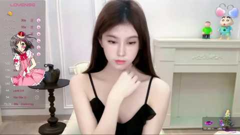 Media: Video of a slender East Asian woman with long black hair in a black dress, sitting indoors with a calendar, toys, and a fireplace in the background.