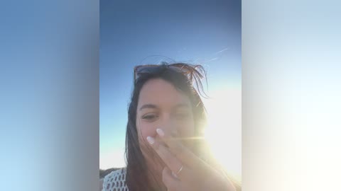 Media: Video of a young woman with long, dark hair, wearing a white knitted sweater, making a peace sign with her fingers, set against a clear blue sky with a sun flare.