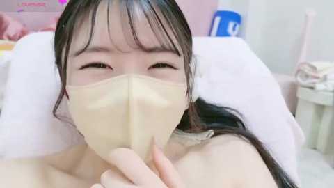 Media: A video of an East Asian woman with long black hair, wearing a beige face mask, lying on a bed in a softly lit room, her expression serene.