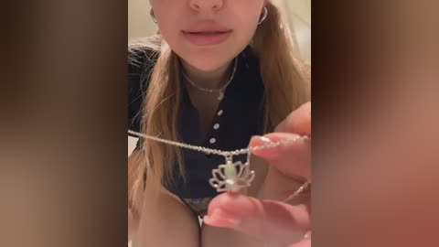 Media: A close-up video of a woman with fair skin and long blonde hair, wearing a black blouse, focusing on a necklace with a silver pendant. The background is blurred.