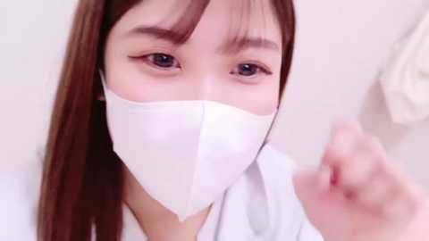 Media: Video of an East Asian woman with straight, shoulder-length brown hair, wearing a white surgical mask, white lab coat, and blue scrubs, holding a syringe in a sterile, clinical setting.