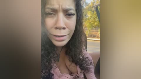 Media: Video of a young woman with curly dark hair and light skin, wearing a pink ribbed sweater, crying with a distressed expression, taken from a car window.