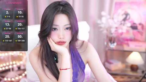 Media: Video of an East Asian woman with long, dark hair, wearing a purple strapless top, sitting pensively on a bed, surrounded by soft, pink, and white decor.