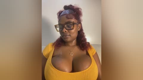 Media: Video of a dark-skinned woman with curly, maroon hair styled in a high bun. She wears large, black-framed glasses, a tight mustard-yellow top revealing ample cleavage, and a delicate necklace. Background is blurred, focusing on her.