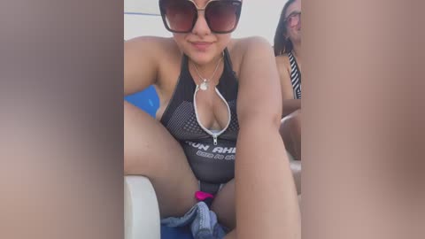 Media: Video of a woman with medium skin tone and dark sunglasses, wearing a black halter swimsuit with \"TUMI\" branding, sitting on a boat with legs spread, revealing a pink bikini bottom, and a friend in the background.