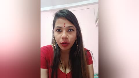 Media: Video of an Indian woman with long dark hair, wearing a red blouse, in a pink room with an air conditioner. She has a nose ring and a dot on her forehead.