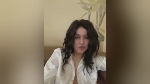 Media: Video of a young woman with wavy, dark hair and light skin, wearing a white shirt with floral patterns, sitting against a beige wall with a potted plant in the background.