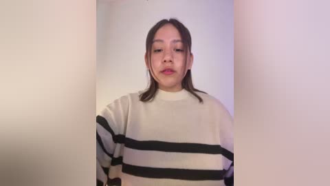 Media: Video of a young Asian woman with straight, shoulder-length brown hair, wearing a white and black striped sweater, standing against a plain white wall.