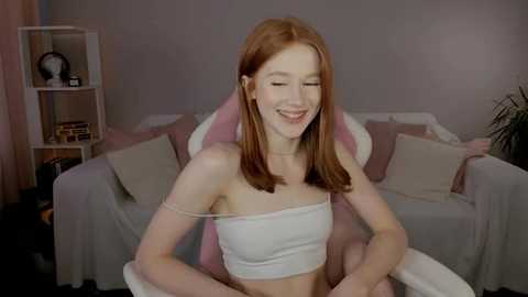 Media: A video of a young, fair-skinned woman with red hair, wearing a white crop top, sitting in a pink gaming chair, smiling in a cozy, pink-themed room.