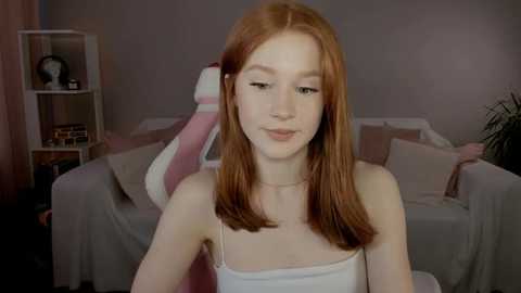 Media: Video of a fair-skinned, slender young woman with straight, shoulder-length auburn hair, wearing a white tank top. Background features a gray couch with light-colored cushions, a white shelving unit with decorative items, and a green potted plant.