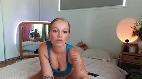 Media: Video of a blonde woman with tattoos on her arms, wearing a blue sports bra, sitting on a bed in a modern bedroom with white walls and a large round lamp.