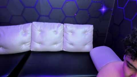 Media: Video of a man with curly hair lying on a white, tufted leather couch, surrounded by hexagonal-patterned walls under purple lighting.