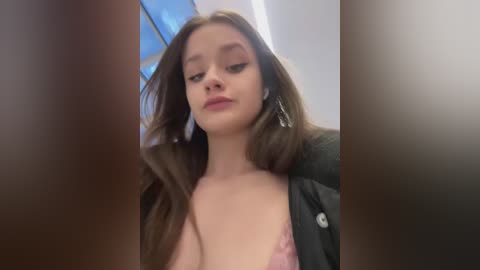 Media: Video of a young Caucasian woman with long, straight brown hair, wearing a black cardigan and a pink lace bra, looking down with a neutral expression. The background features a blue window and a blurred, darkened room.