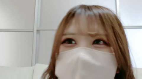 Media: A video of a young Asian woman with light brown hair and fair skin, wearing a white surgical mask, looking down with a serious expression. The background features white tiled walls.