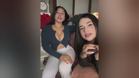 Media: Video of two women with medium skin tones, one with a dark blue crop top and white pants, and the other in a black top, sitting on a couch in a cozy living room.