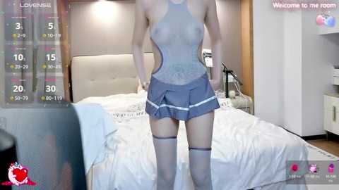 Media: Video of a slender, fair-skinned woman in a sheer, blue lingerie bodysuit and matching mini-skirt, standing in a modern bedroom with a white bed, wooden headboard, and digital clock showing 5:30.