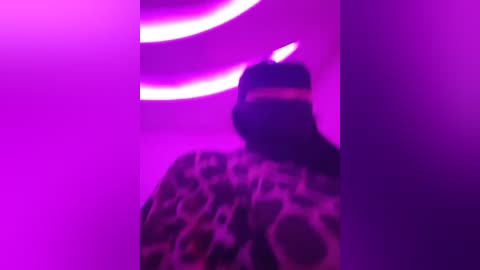 Media: Video of a person with long hair wearing a leopard print hoodie, standing in a dimly lit room with neon pink and purple lights creating a vibrant, surreal atmosphere.