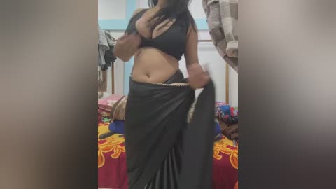 Media: A video of a curvy, light-skinned woman with medium-sized breasts in a black bra and matching skirt, standing in a cluttered bedroom with colorful bedding.