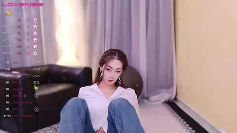 Media: A video of an Asian woman with long brown hair, wearing a white blouse and blue jeans, sitting in a modern living room with a black leather sofa and light-colored curtains.