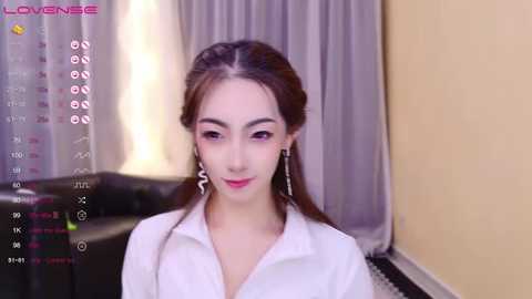 Media: Video of a young East Asian woman with long dark hair, light skin, and wearing a white blouse, standing in a modern, softly lit living room with grey curtains and a black leather couch.