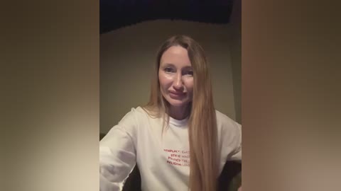 Media: Video of a smiling Caucasian woman with long, straight, light brown hair, wearing a white sweatshirt with red text, in a dimly lit room.