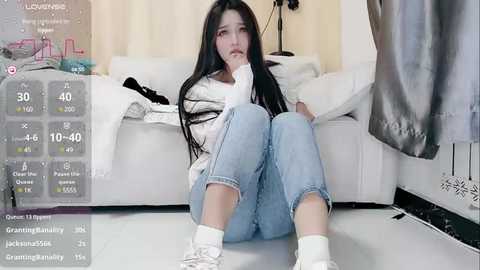 Media: Video of a distressed young woman with long black hair, wearing a white sweater and blue jeans, sitting on a white couch, surrounded by a cluttered living room.