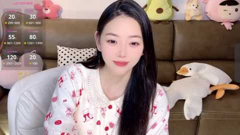 Media: Video of an Asian woman with straight black hair, fair skin, wearing a white pajama shirt with red patterns, sitting on a brown couch with stuffed toys, including a penguin and a frog.