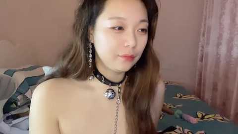 Media: Video of a young Asian woman with long brown hair, wearing a black collar with a bell, silver earrings, and a blue floral-patterned bedspread.