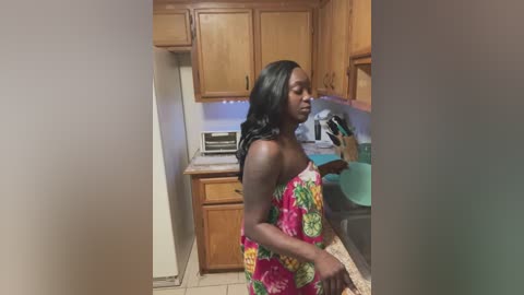 Media: Video of a slender, dark-skinned woman with long black hair, wearing a colorful floral dress, standing in a kitchen with wooden cabinets and beige walls.