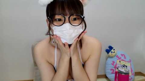 Media: Video of an Asian woman with glasses, wearing bunny ears and a white surgical mask, kneeling in a room with a stuffed toy.